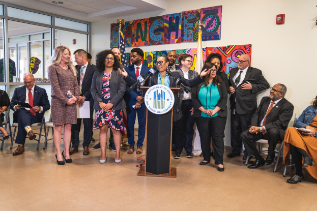 Eine Gruppe von Menschen, die vor einem Podium mit Mikrofon und einer Tafel mit der Aufschrift "Sancho City Councilman Joins City Council" stehen und sitzen, mit Flaggen, Feuerlöscher und Deckenleuchten an der Rückwand.