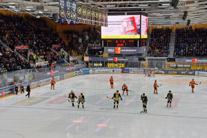 Eine Gruppe von Menschen mit Helmen und Hockeystöcken spielt ein Spiel auf einem Eisstadion mit einem Netz in der Mitte, umgeben von Texttafeln und einem großen Bildschirm im Hintergrund, mit einer Person mit Hut im Vordergrund.