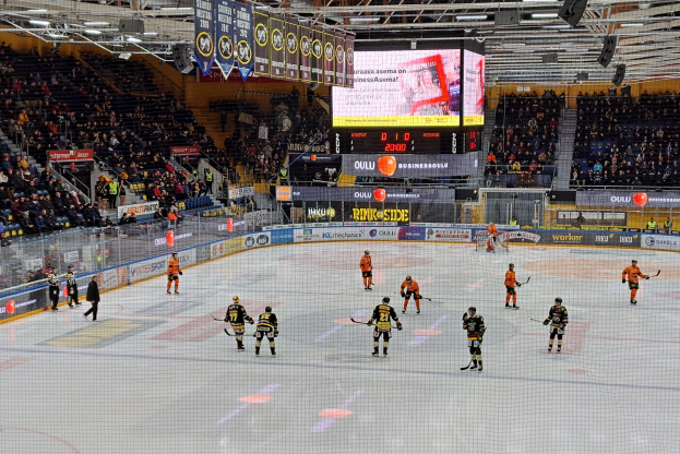 Eine Gruppe von Menschen mit Helmen und Hockeystöcken spielt ein Spiel auf einem Eisstadion mit einem Netz in der Mitte, umgeben von Texttafeln und einem großen Bildschirm im Hintergrund, mit einer Person mit Hut im Vordergrund.