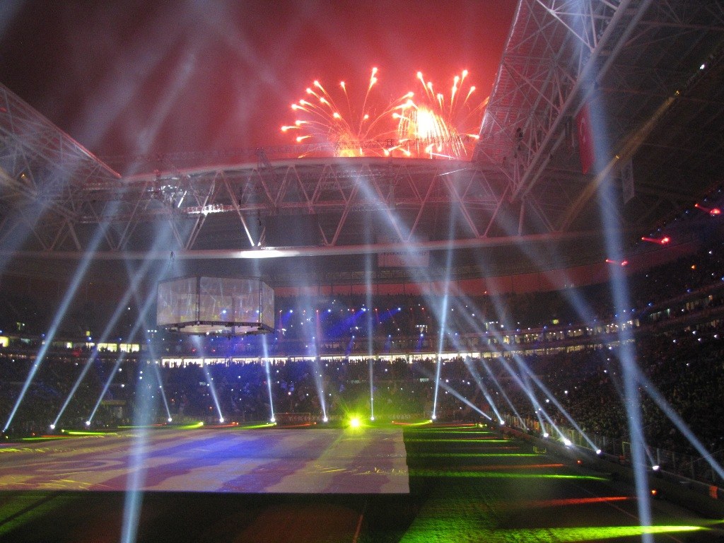 Olympiastadion in London bei Nacht, erleuchtet von hellen Lichtern und Feuerwerken, mit einem großen Bildschirm im Vordergrund und einer Menge drinnen.