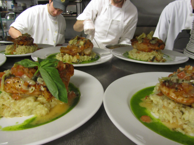 Drei Köche in einer Küche mit Mützen und Handschuhen bereiten Teller mit verschiedenen Speisen zu, mit Gestellen im Hintergrund.