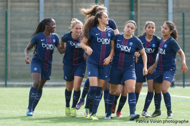 Gruppe junger Frauen beim Fußballspielen auf einem Rasenplatz mit Maschendrahtzaun und einer Wand im Hintergrund, beschriftet mit "Paris Saint-Germain Frauenfußball" in der rechten unteren Ecke.