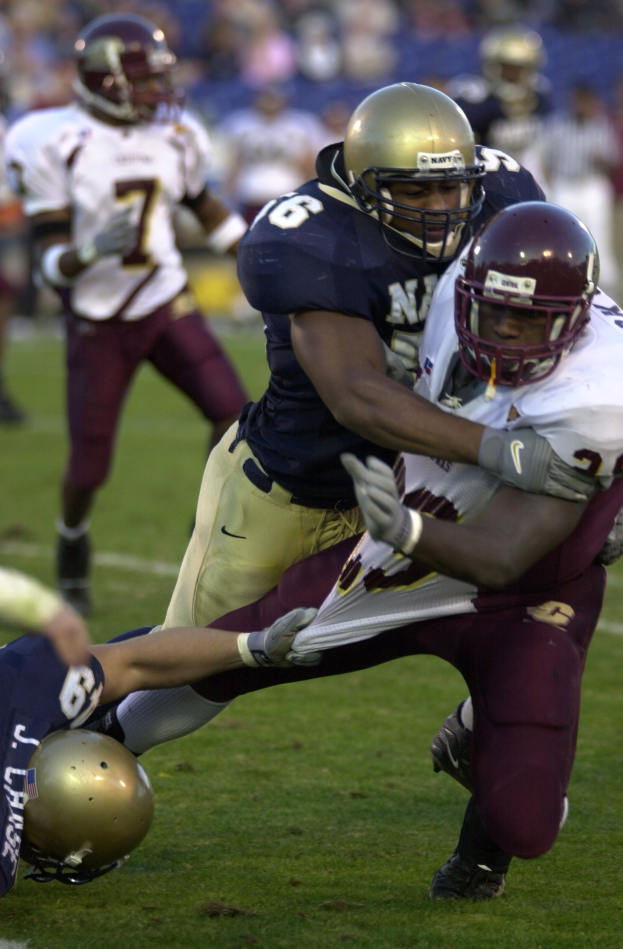 Zwei Fußballspieler in Sportkleidung und Helmen, einer den anderen auf dem Feld tackelnd, mit anderen Spielern und Zuschauern im Hintergrund.