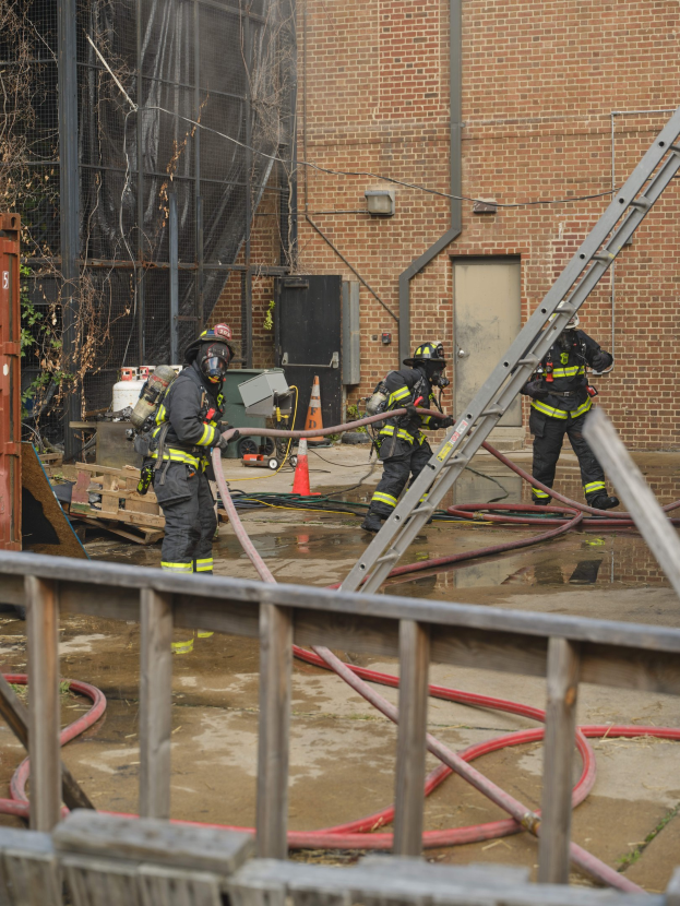 Feuerwehrleute in Helmen arbeiten daran, ein Gebäude Feuer zu löschen, umgeben von Ausrüstung, einem Zaun und Hintergrundelementen einschließlich eines Baumes und des Himmels.