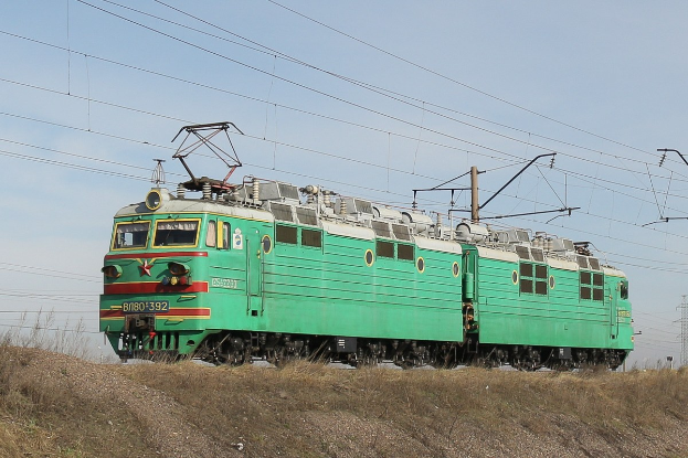 Ein grüner Elektrolokomotive fährt auf Bahnschienen durch ein grünes Feld mit Strommasten und einem Turm im Hintergrund unter einem klaren blauen Himmel.