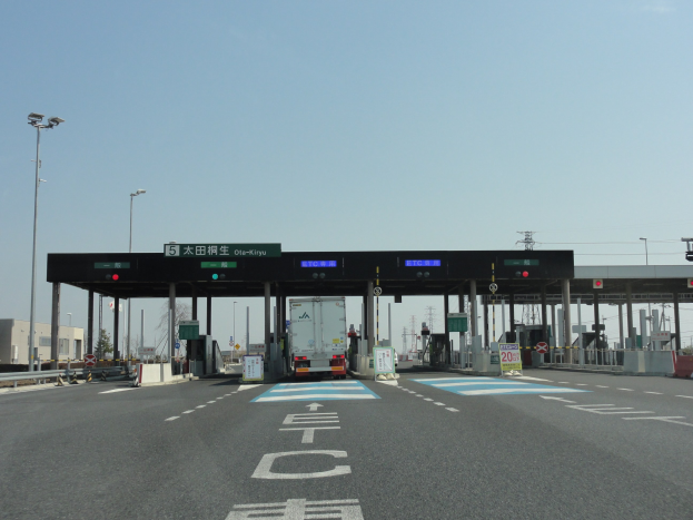 Mautstelle mit einem vor ihr geparkten Lkw auf einer stra├čenrandseitig mit wei├čen und blauen Markierungen versehenen Stra├če mit Laternenmasten, Schildern und Geb├Ąuden im Hintergrund bei einem klaren blauen Himmel.