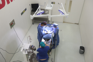 Chirurgen führen einen Eingriff an einem Patienten in einem Operationssaal durch, mit medizinischer Ausrüstung an der Wand und einem Objekt auf dem Boden rechts.