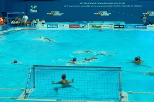 Eine Gruppe von Menschen, die Wasserball in einem Pool mit einem Netz spielen, mit Zuschauern im Hintergrund und Bildschirmen, die die 2015 FINA Weltmeisterschaft in Kazan zeigen.