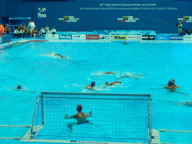 Eine Gruppe von Menschen, die Wasserball in einem Pool mit einem Netz spielen, mit Zuschauern im Hintergrund und Bildschirmen, die die 2015 FINA Weltmeisterschaft in Kazan zeigen.