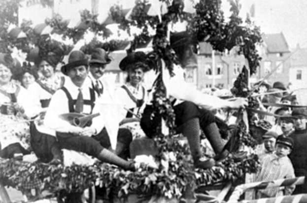 Eine Gruppe von Menschen in traditioneller bayrischer Tracht fährt auf einem mit Laub geschmückten Festwagen mit, einige spielen Musikinstrumente, im Hintergrund sind Gebäude und ein klarer Himmel zu sehen, dargestellt in Schwarz-Weiß.