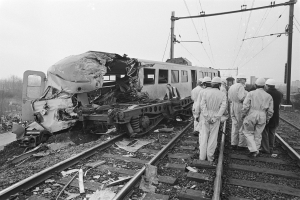 Eine Schwarz-Weiß-Szene eines Zugunglücks mit Menschen in Helmen, die sich darum versammeln, Strommasten, Bäume, Gebäude und einen klaren Himmel im Hintergrund.