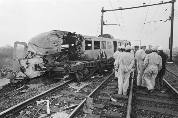 Eine Schwarz-Weiß-Szene eines Zugunglücks mit Menschen in Helmen, die sich darum versammeln, Strommasten, Bäume, Gebäude und einen klaren Himmel im Hintergrund.