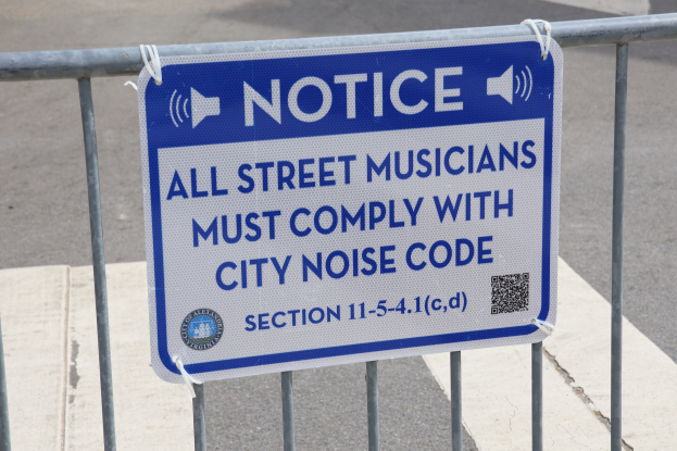 Anschlagtafel mit einem Schild, auf dem "Anschlag: Alle Straßenmusiker müssen die Stadtlärmverordnung einhalten" steht, an einem Zaun befestigt, mit einer Straße im Hintergrund.