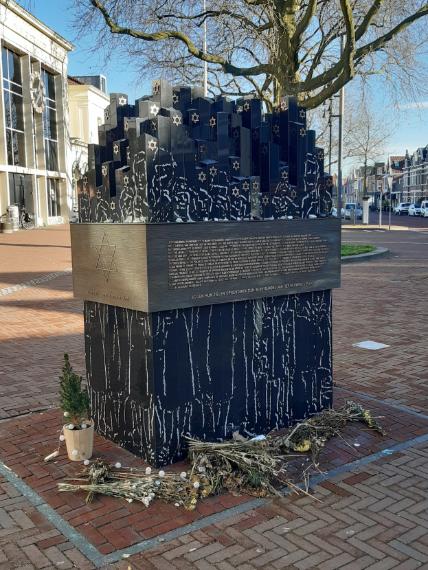 Denkmal in einem Stadtplatz, das an Holocaust-Opfer erinnert, mit einem Baum davor, einer Topfpflanze, verstreuten trockenen Blättern und Gebäuden, Fahrzeugen und Bäumen im Hintergrund.
