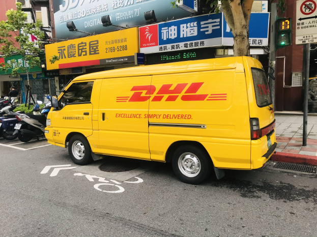 Ein gelber DHL-Lieferwagen parkt auf einer Straße neben Gebäuden, Bäumen, Schildern, Pfählen, Verkehrsampeln und einem Fußgänger auf dem Gehweg.
