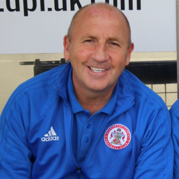 Zwei Männer in einem Raum, einer trägt eine blaue Jacke und lächelt neben einem anderen Mann, im Hintergrund eine Wandtafel.