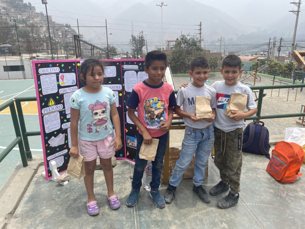 Gruppe von Kindern mit Papiertüten auf einem Basketballfeld mit Tüten auf dem Boden, eine Tafel mit Text links und Hintergrund-Elemente wie Zaun, Pfosten, Straßenlaternen, Bäume, Gebäude, Hügel und Himmel.