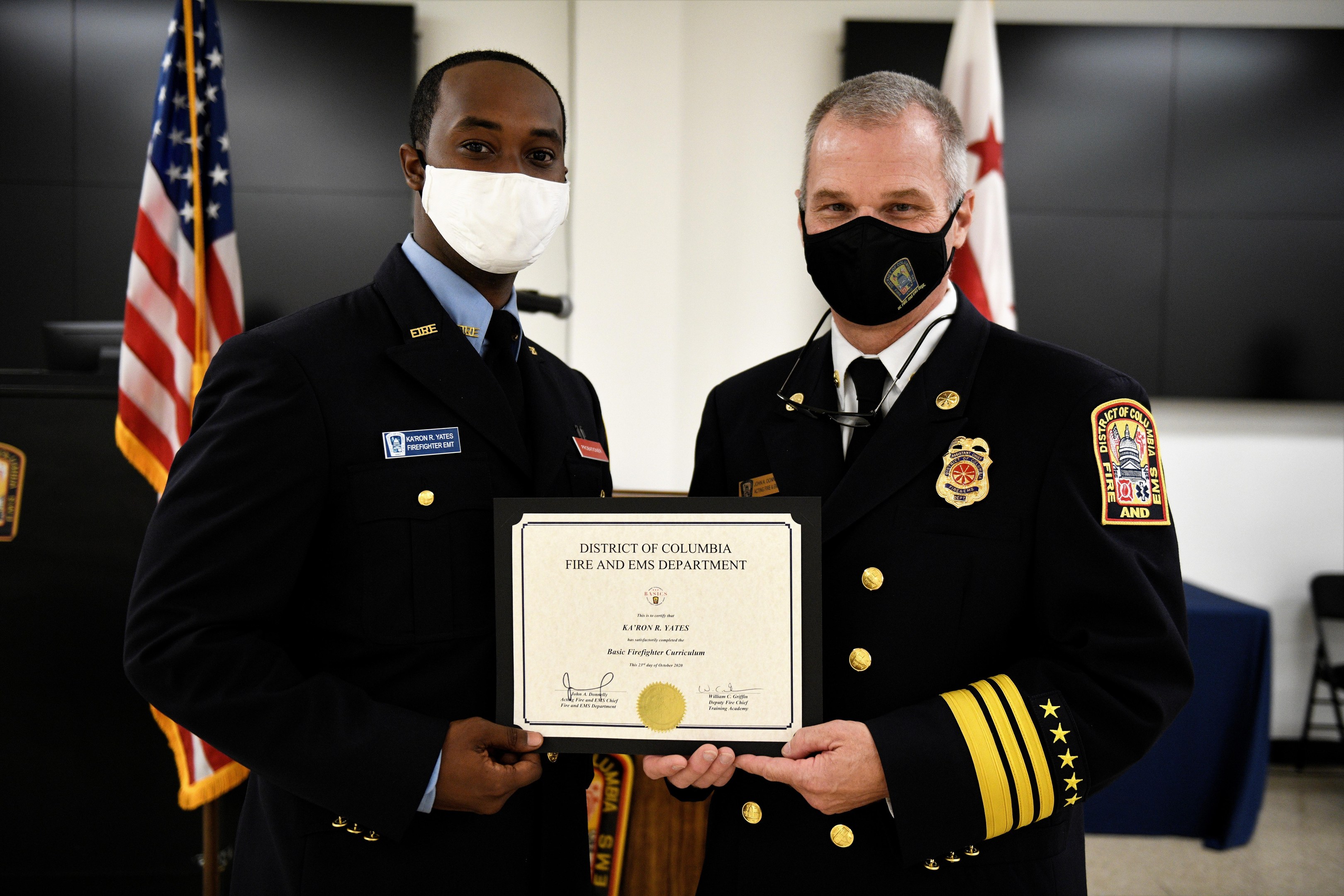 Zwei Männer in Feuerwehrausstattung stehen nebeneinander und tragen Masken sowie ein Zertifikat; im Hintergrund sind Flaggen und Möbel zu sehen; der Mann links ist der Feuerwehrchef des District of Columbia und erhält eine Auszeichnung.
