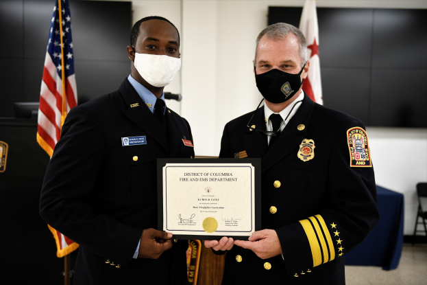 Zwei Männer in Feuerwehrausstattung stehen nebeneinander und tragen Masken sowie ein Zertifikat; im Hintergrund sind Flaggen und Möbel zu sehen; der Mann links ist der Feuerwehrchef des District of Columbia und erhält eine Auszeichnung.