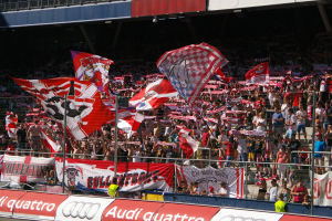 Große Menschenmenge in einem Stadion mit Fahnen, Bannern, einem Metallzaun, einem Schild, einem Anzeigebildschirm und Deckenleuchten.