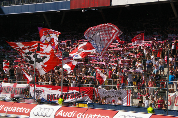 Große Menschenmenge in einem Stadion mit Fahnen, Bannern, einem Metallzaun, einem Schild, einem Anzeigebildschirm und Deckenleuchten.