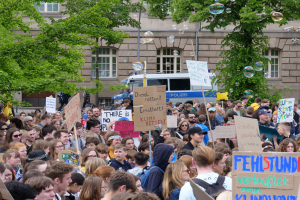 Eine große Gruppe von Menschen mit Schildern vor einem Gebäude mit Fenstern, Bäumen und einem Fahrzeug im Hintergrund, mit Blasen in der Luft, die an einer Klimademonstration in Berlin teilnehmen.