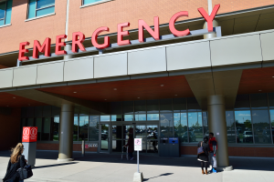 Eine Gruppe von Menschen, die vor einem gläsernen Notrufgebäude mit Säulen, einem Namenbrett, Schildern auf Pfählen und einem Mülleimer gehen.