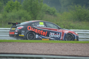 Ein Auto auf einer Rennstrecke umgeben von Gras, einem Geländer und Bäumen im Hintergrund mit Text und Zahlen auf dem Auto.