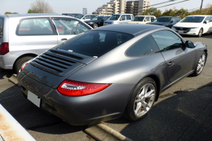 Ein silberner Porsche 911 auf einem Parkplatz mit anderen Autos, Gebäuden, Bäumen, Polen, Drähten und einem klaren blauen Himmel.