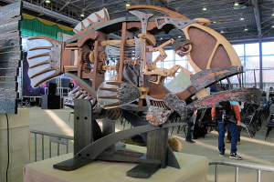 Große Metallfischskulptur in einem Innenraum mit Zuschauern, umgeben von Stühlen, einem Metallzaun, einer Fahne, einem Lautsprecher, Vorhängen und Deckenleuchten.