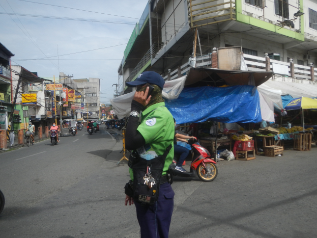 Ein Polizist in grüner und blauer Uniform und einer blauen Mütze steht an der Seite einer Straße und spricht in ein Handy, während im Hintergrund Fahrzeuge fahren, rechts von ihm Stände mit verschiedenen Gegenständen und ein bewölkter Himmel zu sehen sind.
