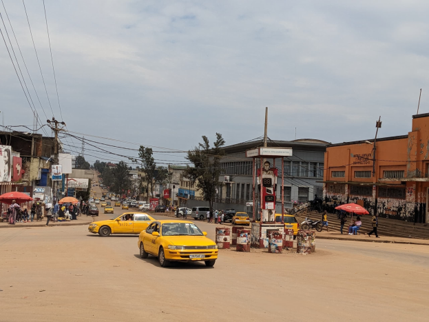 Ein gelbes Taxi fährt eine belebte Stadtstraße entlang, gesäumt von Gebäuden, während Fußgänger mit Regenschirmen auf dem Gehweg unter bewölktem Himmel gehen, mit Strommasten und Drähten im Hintergrund.