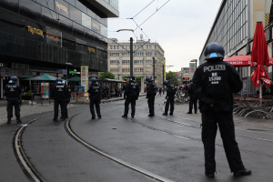 Gruppe von Polizisten in schwarzen Uniformen und Helmen auf einer Straße mit Gebäuden, Glasfenstern und verstreuter Außenmöbel unter einem klaren Himmel.