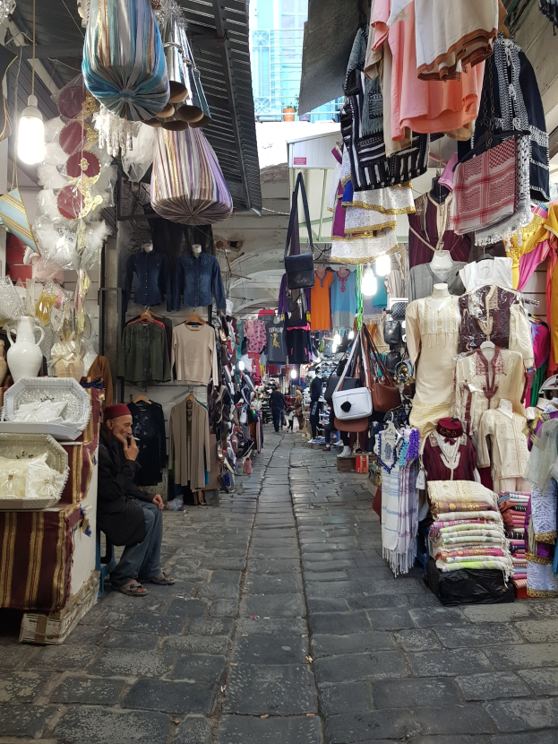 Ein belebter Markt in der Altstadt von Jerusalem, mit Menschen, die an Ständen mit Kleidung, Taschen, Lampen und anderen Gegenständen vorbeischlendern, und einem Gebäude im Hintergrund.