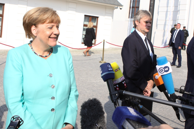 Bundeskanzlerin Angela Merkel beim Sprechen mit Reportern vor einem Gebäude mit Fenstern und einem bewölkten Himmel, umgeben von einer Gruppe von Menschen, einige halten Mikrofonstative.