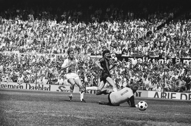Schwarz-weiß-Foto von Männern, die auf einem Feld Fußball spielen, mit Zuschauern im Hintergrund und einer Bundesliga 1978-79 Ajax 1-1 Hannover 96-Fahne.