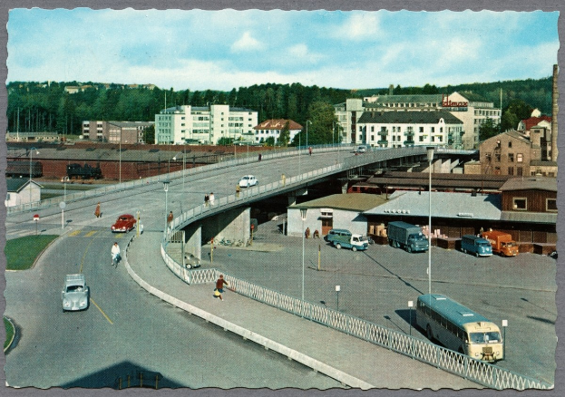 Altes Schwarz-Weiß-Foto einer Stadtstraße mit fahrenden Autos und Bussen, Fußgängern auf einer Brücke, Laternenmasten, mehrstöckigen Gebäuden mit Fenstern, Bäumen und einem bewölkten Himmel.