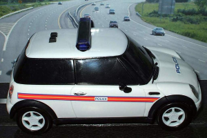 Ein weißes Polizeiauto steht im Vordergrund mit mehreren fahrenden Autos auf der Straße dahinter, umgeben von Gras und Bäumen in einer ländlichen Umgebung.