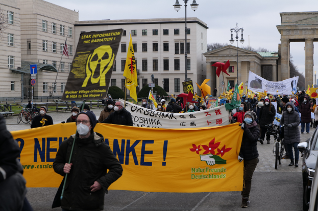 Große Gruppe von Menschen bei einer Straßendemonstration gegen Atomkraft in Deutschland, mit Transparenten und Fahnen, Fahrzeugen auf der rechten Seite, Gebäuden und Bäumen im Hintergrund und einem Tor in der Ferne.