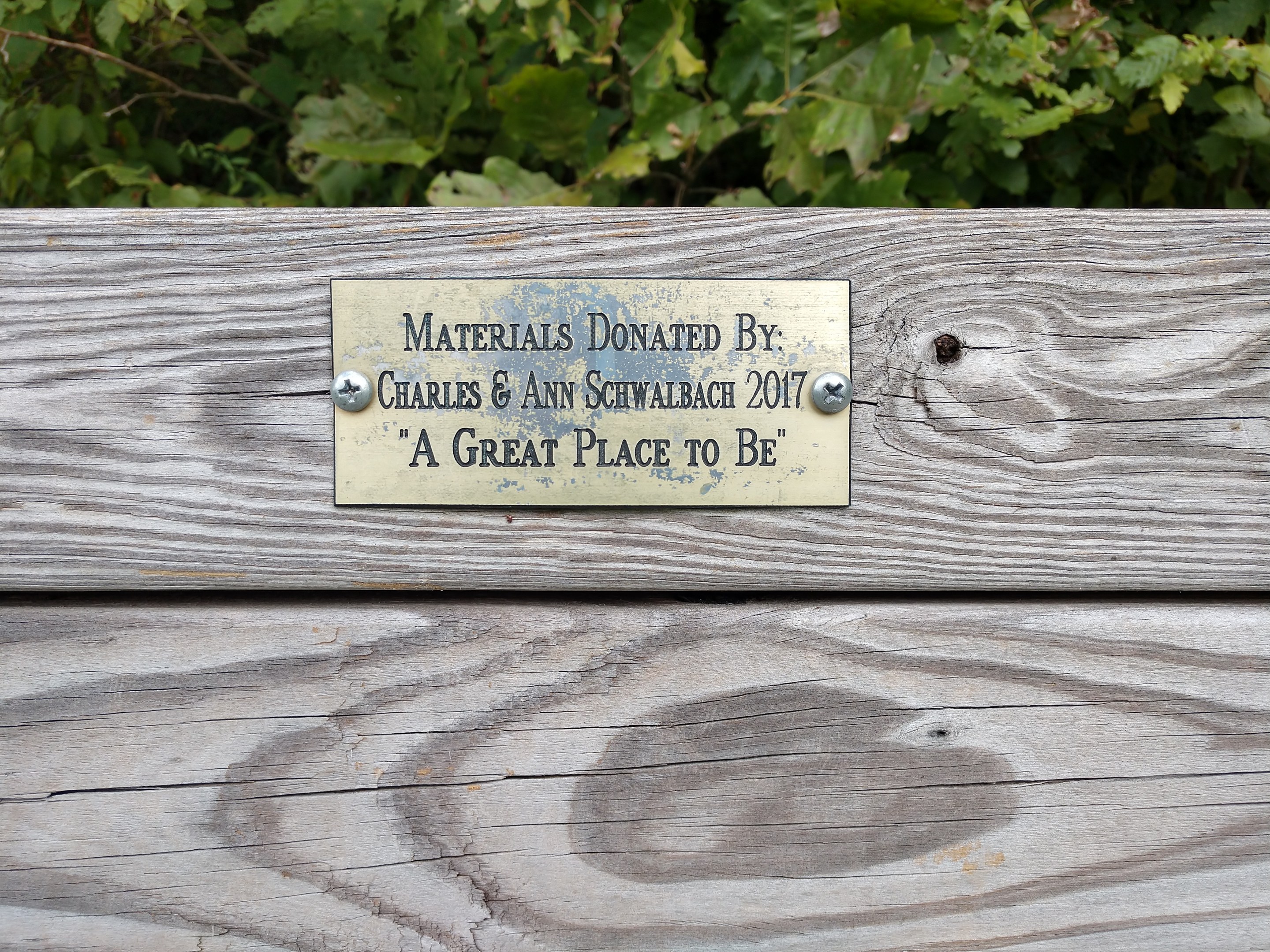 Holzbank mit einer Plakette, auf der "Materialien gespendet von Charles & Ann Schwabach 2017: Ein schöner Ort zu sein" steht, in einem Garten mit üppiger grüner Vegetation.