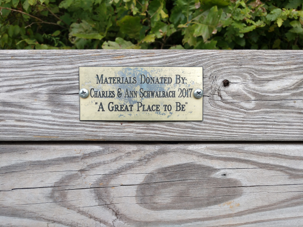 Holzbank mit einer Plakette, auf der "Materialien gespendet von Charles & Ann Schwabach 2017: Ein schöner Ort zu sein" steht, in einem Garten mit üppiger grüner Vegetation.