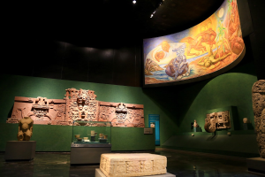 Ausstellungsraum im Anthropologischen Museum von Mexiko-Stadt, der verschiedene Kunstwerke zeigt, darunter Skulpturen, Tischgegenstände, eine Texttafel, eine Wandmalerei und eine Deckenbeleuchtung.