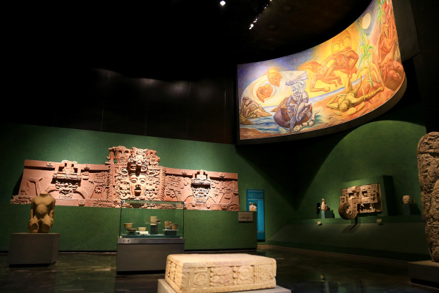 Ausstellungsraum im Anthropologischen Museum von Mexiko-Stadt, der verschiedene Kunstwerke zeigt, darunter Skulpturen, Tischgegenstände, eine Texttafel, eine Wandmalerei und eine Deckenbeleuchtung.