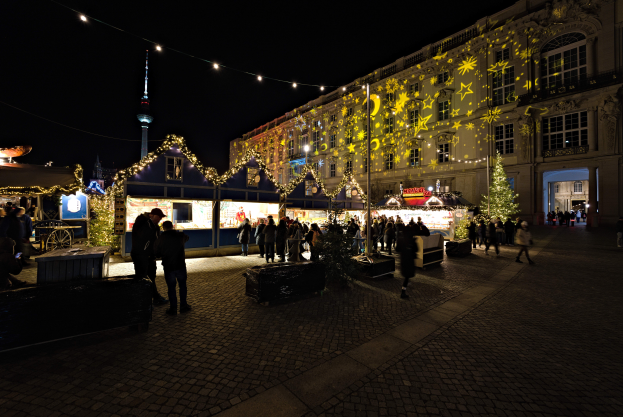 Ein lebendiger Weihnachtsmarkt in Berlin, Deutschland, mit Menschen um beleuchtete und geschmückte Stände, vor Gebäuden mit Fenstern unter einem dunklen Himmel.