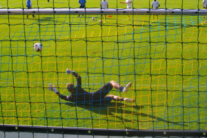 Ein Fussballer taucht ab, um einen Ball vor dem Tor zu halten, mit einem Netz im Vordergrund und einigen Menschen im Hintergrund, während mehrere Fussbälle auf dem Boden verstreut sind.