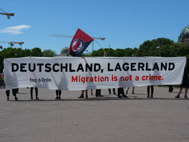 Eine Gruppe von Menschen mit einem Banner, auf dem 'Deutschland, Lagerland Migration ist kein Verbrechen' steht, und einer Flagge, im Hintergrund Straßenlaternen, Bäume, Kräne, ein Gebäude und ein bewölkter Himmel.
