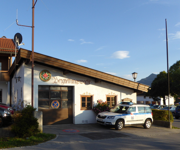 Polizeiauto vor einem Gebäude mit Fenstern und Türen geparkt, umgeben von Grünflächen einschließlich blühender Pflanzen, Gras und Bäumen, mit Straßeninfrastruktur wie Pfählen und einem Schild sichtbar unter einem bewölkten Himmel.