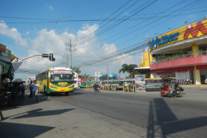 Eine belebte Stadtstraße mit Fahrzeugen wie einem Bus und Motorrädern, Fußgänger auf Gehwegen, Verkehrsampeln, Strommasten, Gebäude mit Namensschildern, Bäume und ein bewölkter Himmel.