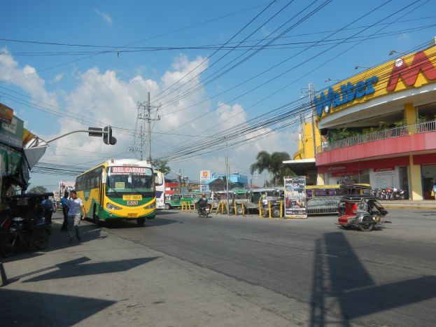 Eine belebte Stadtstraße mit Fahrzeugen wie einem Bus und Motorrädern, Fußgänger auf Gehwegen, Verkehrsampeln, Strommasten, Gebäude mit Namensschildern, Bäume und ein bewölkter Himmel.