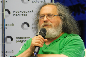 Ein Mann mit langen Haaren und Brille sitzt auf einem Stuhl und hält ein Mikrofon in der Hand, während er ein grünes T-Shirt trägt und ein Banner im Hintergrund darauf hinweist, dass er Psycholinguist ist.